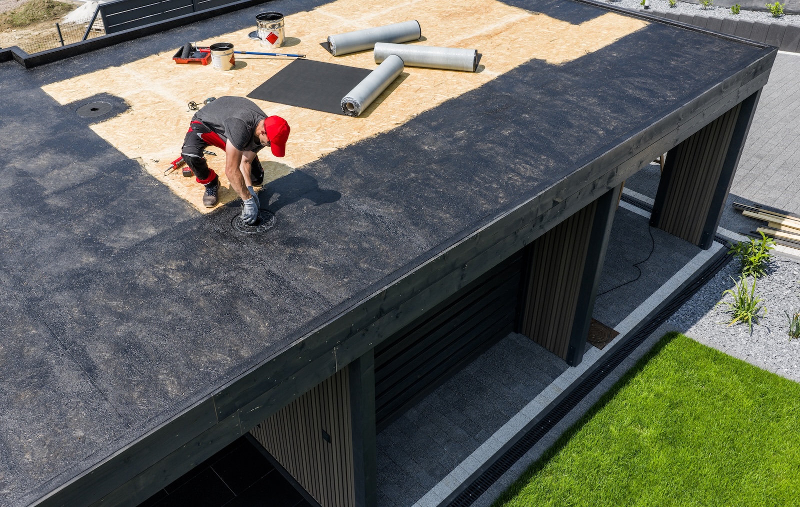 A construction worker is installing asphalt rolling roofing material on a contemporary home under clear skies, surrounded by tools and prepared materials for the roofing project.